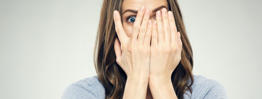 Woman covering up her entire face by opening her fingers to see through with one eye, looking fearful